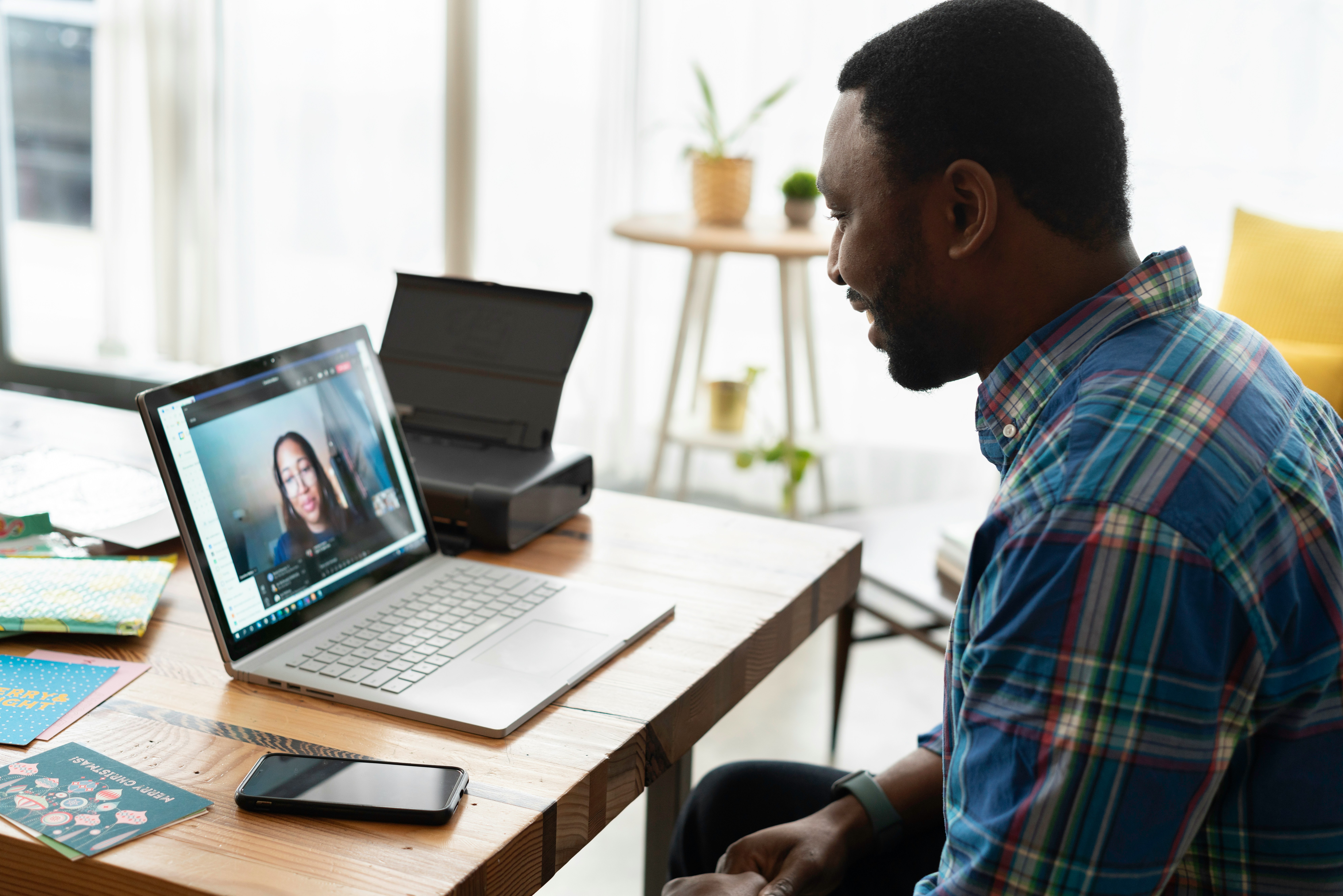 A man in a plaid shirt is sitting at a wooden desk, engaged in a video call on his laptop. The screen displays a woman talking, suggesting a professional or personal online meeting. The desk also features a smartphone, some colorful documents, and a printer in the background. The room is well-lit with natural light, creating a comfortable and productive workspace.