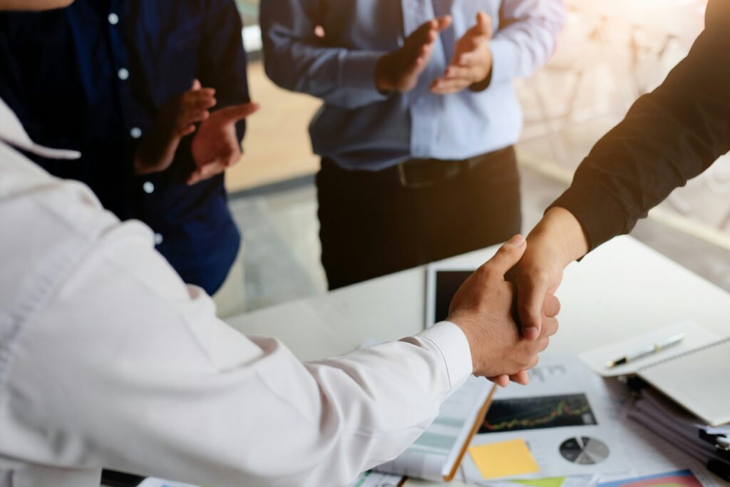 A close-up image of a professional setting where two individuals are shaking hands, signifying a successful agreement or partnership. In the background, other individuals are clapping, suggesting a congratulatory or celebratory atmosphere. The scene includes a desk with documents, charts, and a tablet, indicating a business environment. The overall mood is positive and collaborative.